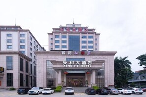 Un hotel amb una entrada destacada, diverses plantes i un aparcament amb diversos cotxes.