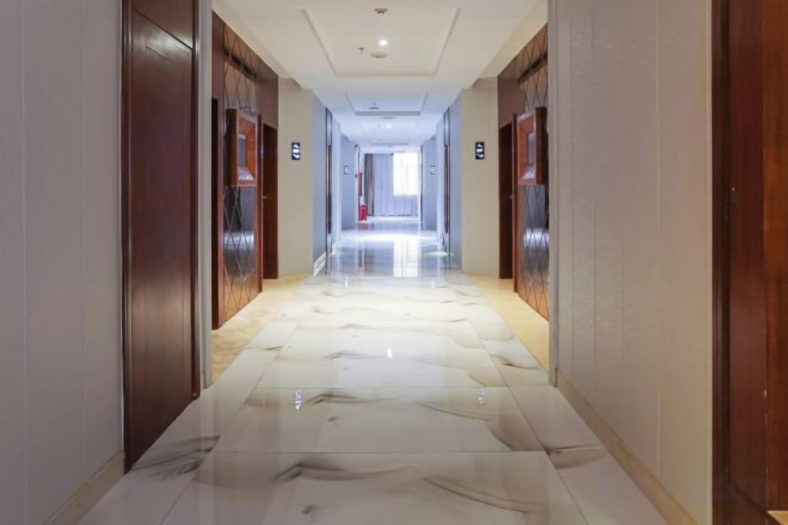 Un couloir d’hôtel avec un sol en marbre, des portes en bois et une lumière vive au bout.