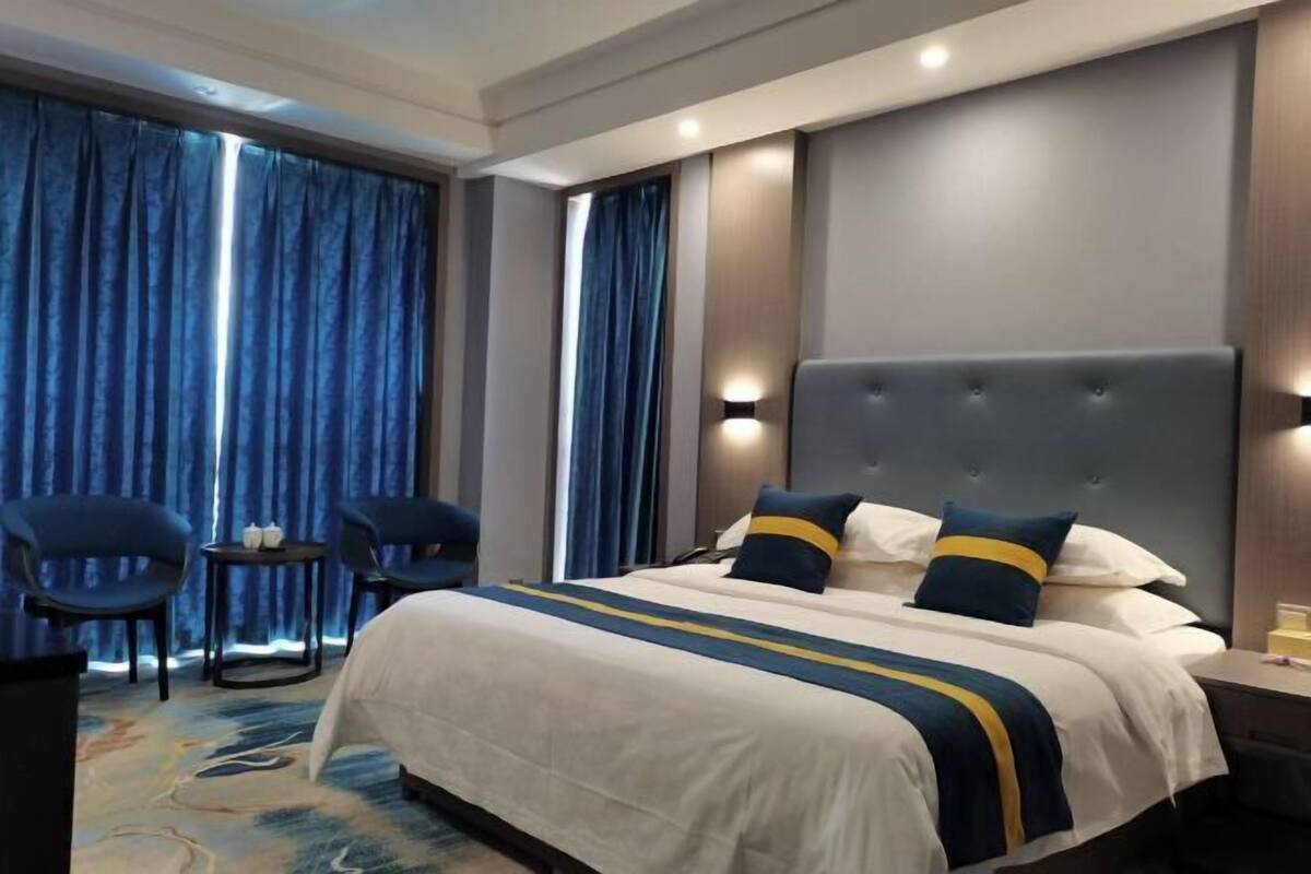 Hotellihuone, jossa on suuri sänky, kaksi tuolia, pieni pöytä ja suuri ikkuna, jossa on verhot.
