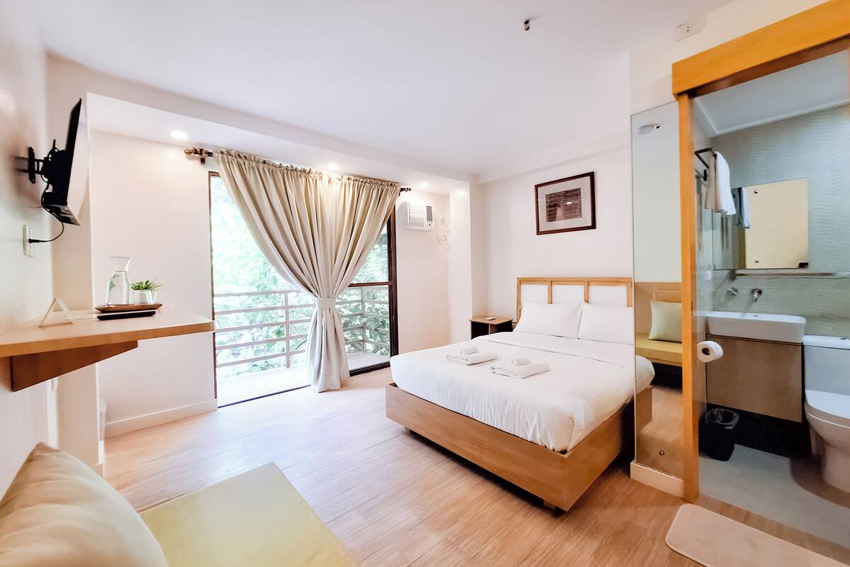 Un dormitorio moderno con cama de madera, una mesita de noche, un tocador, un baño con espejo y lavabo, y un balcón con cortinas.