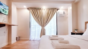 A modern hotel room with a large bed, a flat-screen TV mounted on the wall, a small bedside table with a lamp, and a window with sheer curtains. - Areca Boracay Bed & Breakfast (Boracay Island)