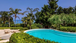 A swimming pool surrounded by lush greenery and palm trees, with a clear view of the ocean in the background. - The Sicily Lobster House (naso)