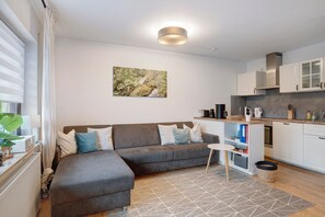A modern living room with a sectional sofa, a small table, and a kitchen area with white cabinets and a built-in stove.