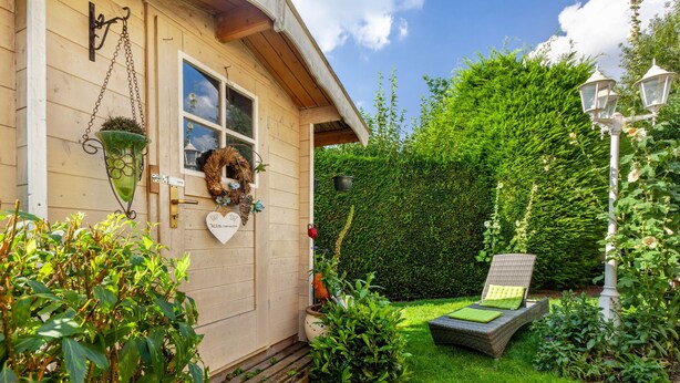 A wooden house with a front door, a hanging flower basket, a small garden with a chair, and a hedge. - Apartment 'Haus Ute' with Private Terrace, Private Garden and Wi-Fi (Arnsberg)