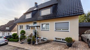 A two-story house with a tiled roof, a paved driveway, and a small patio area with outdoor furniture. - Apartment 'Haus Ute' with Private Terrace, Private Garden and Wi-Fi (Arnsberg)