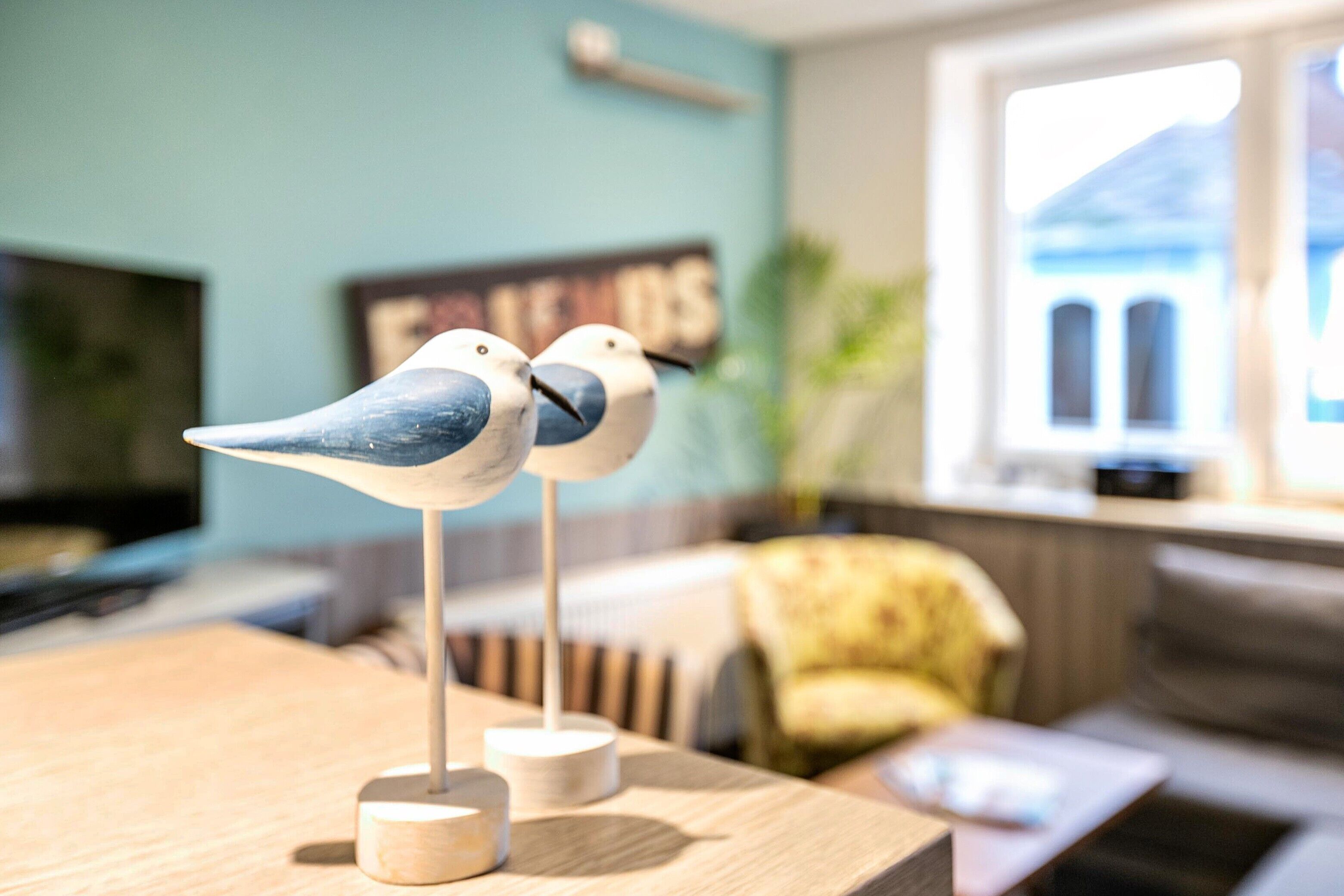 Une pièce avec une table en bois, deux décorations en forme d’oiseau, un téléviseur, un fauteuil jaune et une fenêtre donnant sur un immeuble.