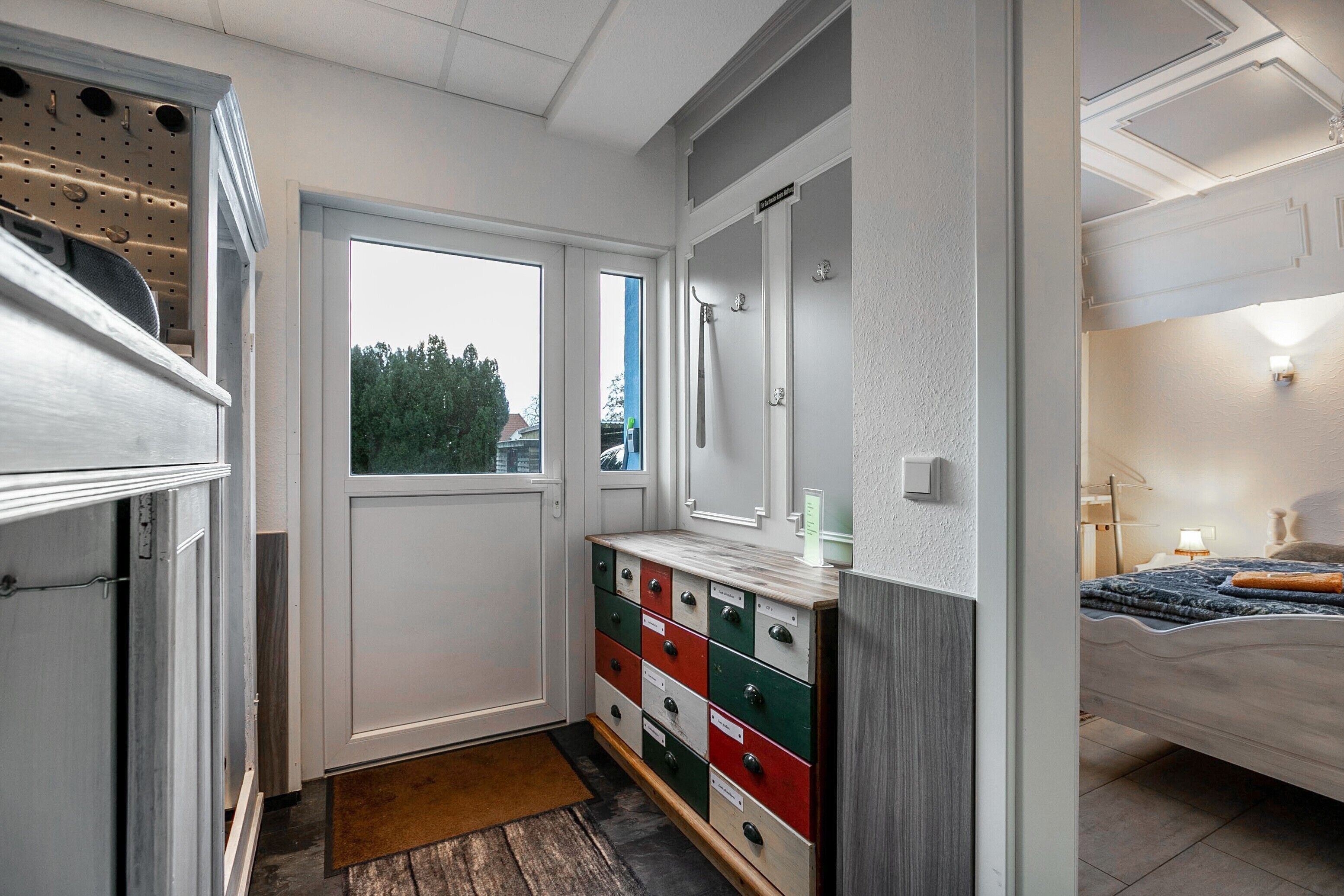 A compact room with a bed, a dresser with drawers, a mirror, a door leading to another room, and a window with a view of trees.