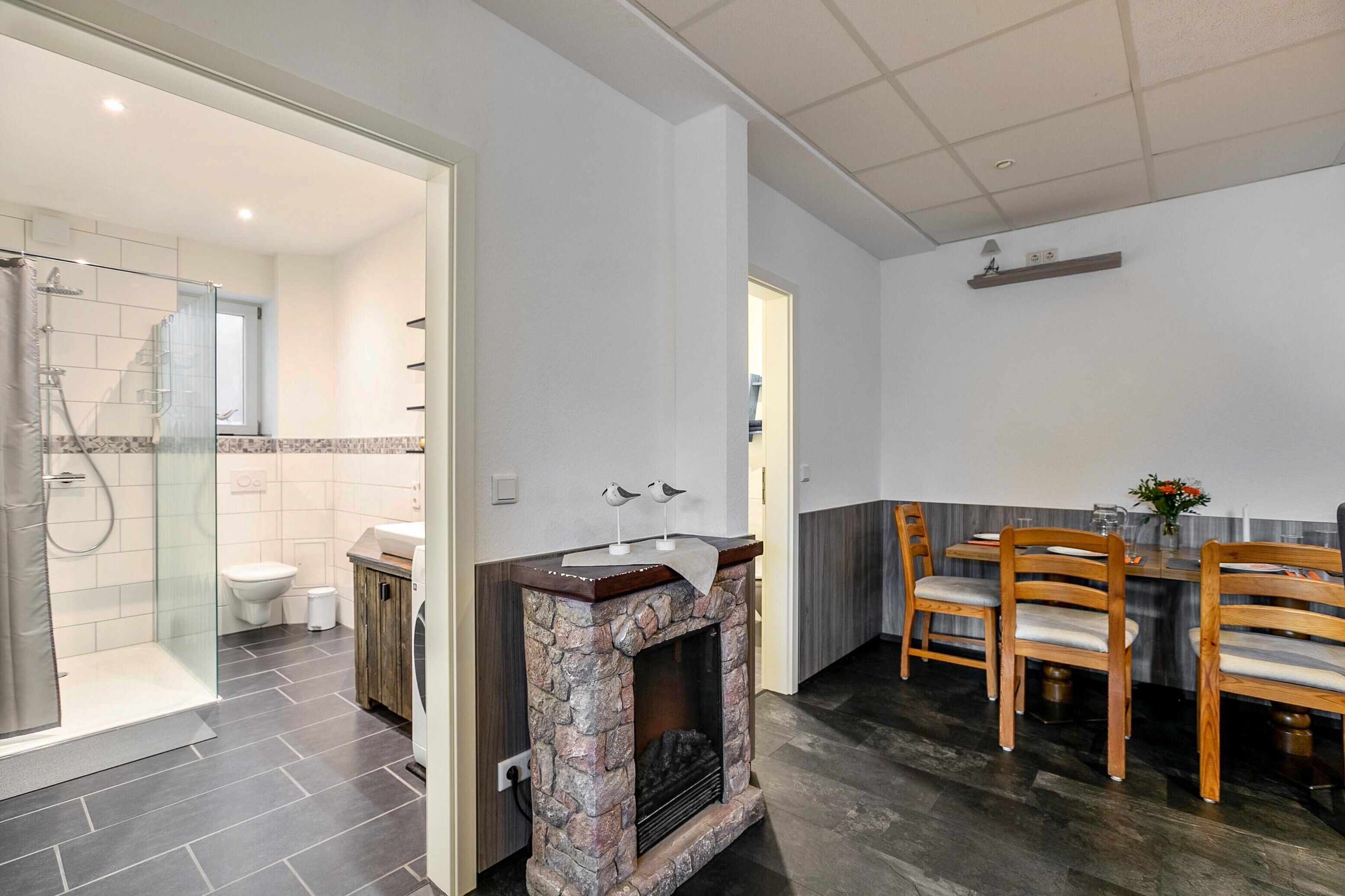 Une salle de bain moderne avec une douche, des toilettes et un lavabo. On y trouve également un coin repas avec des chaises et une table en bois, ainsi qu’une cheminée avec un manteau en pierre.
