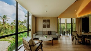 A spacious living area with a sofa, coffee table, and dining table, large windows offering a view of greenery and palm trees. - Sunyi Ubud (Payangan)