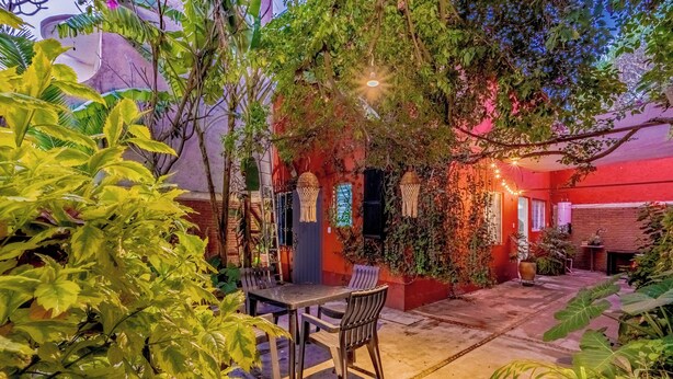 A patio area with a table and chairs, surrounded by lush greenery and a red building facade. - Residencia en la ciudad de Oaxaca (Oaxaca de Juárez)