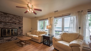 A living room with a fireplace, two sofas, a wooden coffee table, and a ceiling fan. - Rustic, Cozy Lakefront Cabin with Private Beach on Upper Chateaugay Lake. (Lyon Mountain)