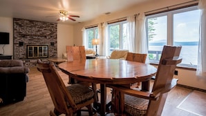 A spacious living room with a large wooden table and chairs, a fireplace, and a view of the lake. - Rustic, Cozy Lakefront Cabin with Private Beach on Upper Chateaugay Lake. (Lyon Mountain)