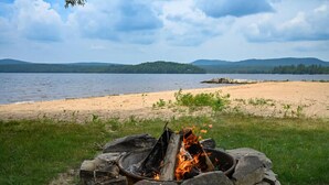 A campfire in a stone fire pit by a lakeside with a view of distant hills. - Rustic, Cozy Lakefront Cabin with Private Beach on Upper Chateaugay Lake. (Lyon Mountain)