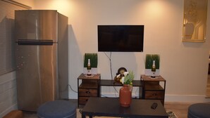 A modern living room with a stainless steel refrigerator, a flat-screen TV mounted on the wall, two wooden side tables with decorative vases, and a central coffee table.