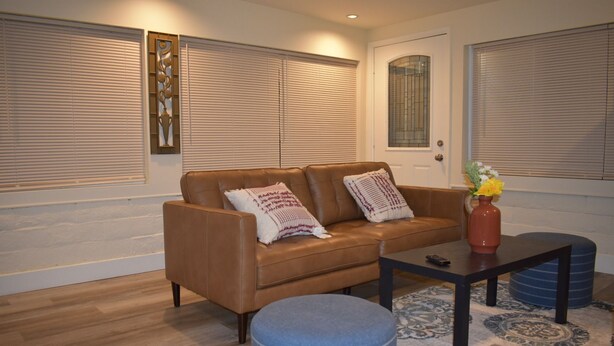 A living room with a brown leather sofa, two patterned throw pillows, a small table with a vase of flowers, and a blue ottoman.