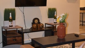 A living room with a flat-screen TV mounted on the wall, a brown sofa, a coffee table, and decorative items including a vase with flowers and a bottle of wine.