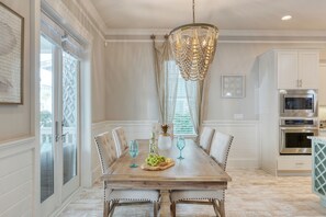 A dining area with a wooden table, white upholstered chairs, a chandelier, and a kitchen with stainless steel appliances. - Shine on the Shore by AvantStay | Pool + Golf Cart (Inlet Beach)