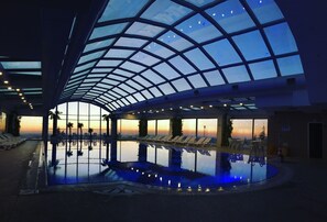 An indoor swimming pool with a glass ceiling, lounge chairs, and a sunset view.
