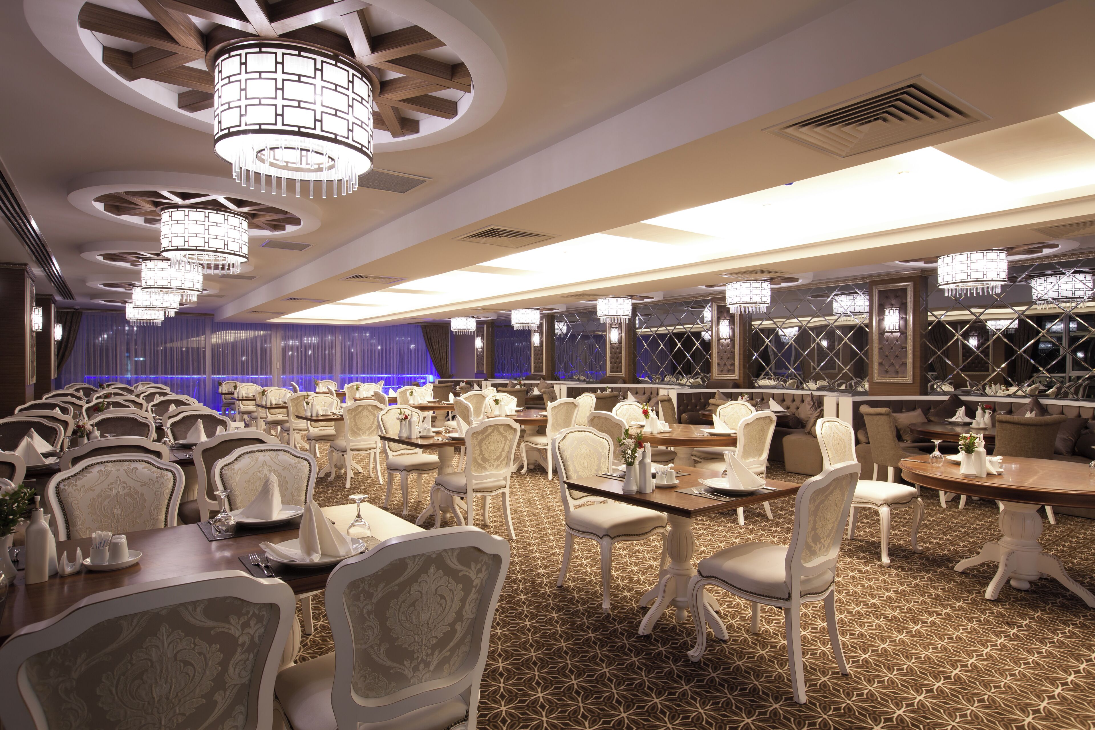 A banquet hall with round tables and chairs, chandeliers, and a stage with blue curtains.