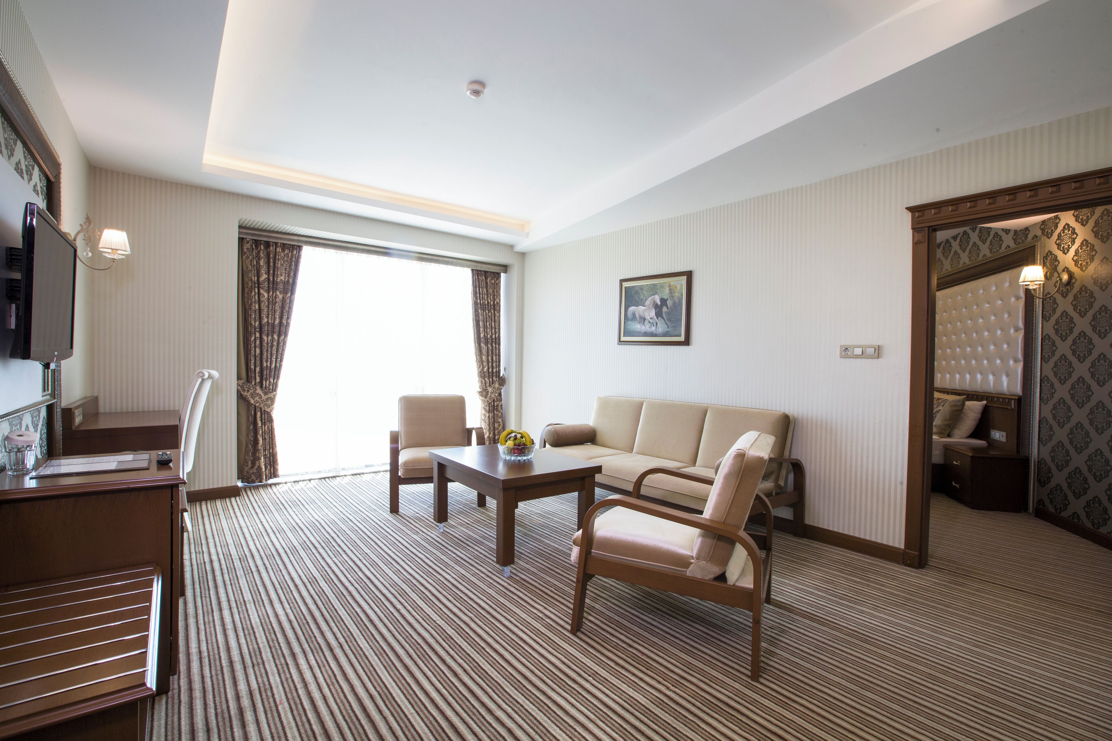 Quarto de hotel com sofá, duas poltronas, mesa de centro, televisão e uma grande janela com cortinas.