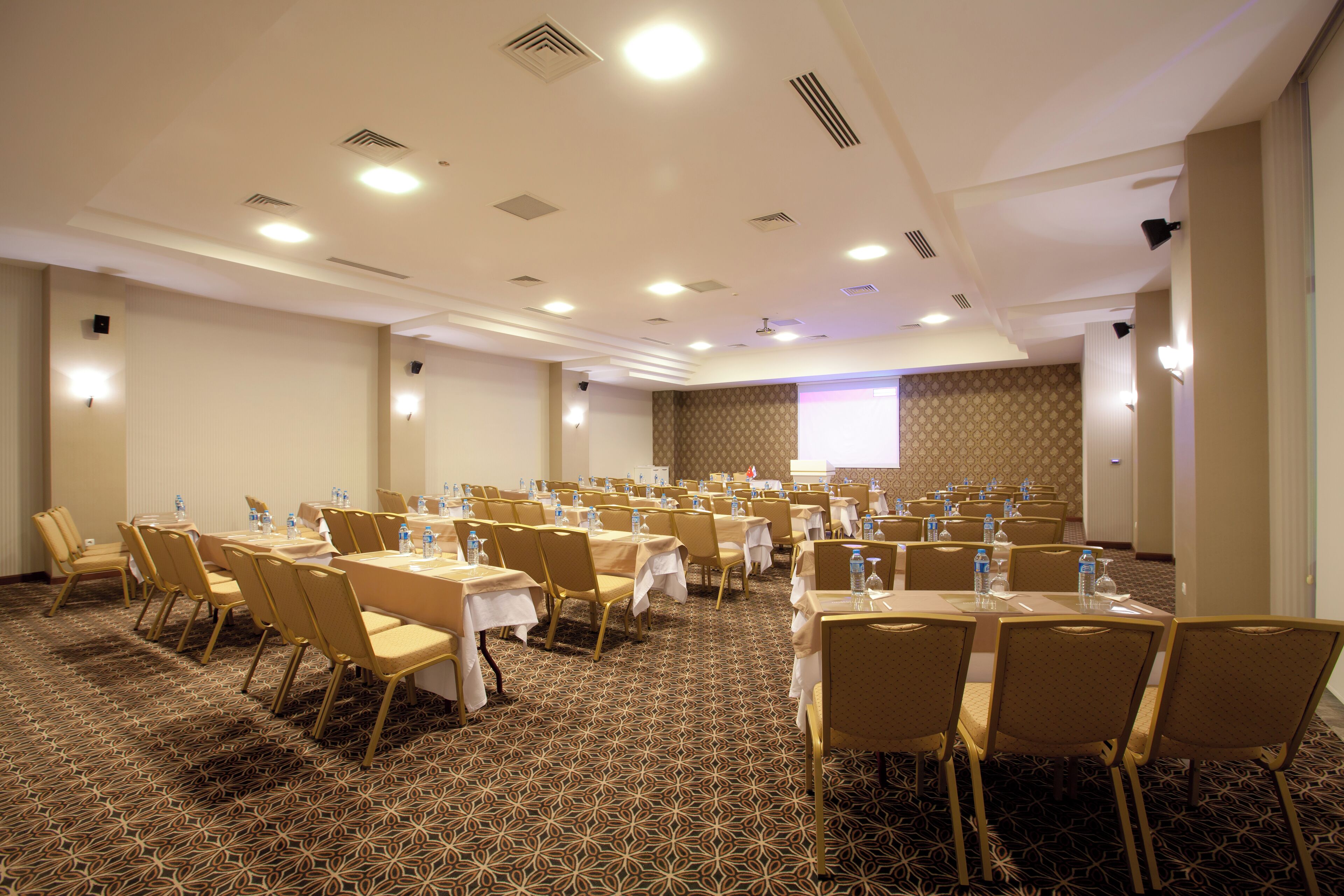 A conference room with tables and chairs, equipped with water bottles and a projector screen.