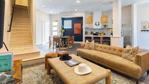 A modern living room with a sofa, ottoman, and a dining area with chairs and a table. A staircase leads to another level. - 10 Min to Downtown | The Outrider 4BR Suite (Nashville)