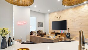 A modern living room with a flat-screen TV, wooden wall paneling, a neon sign, and a kitchen area with a sink and fruit bowl. - 10 Min to Downtown | The Outrider 4BR Suite (Nashville)