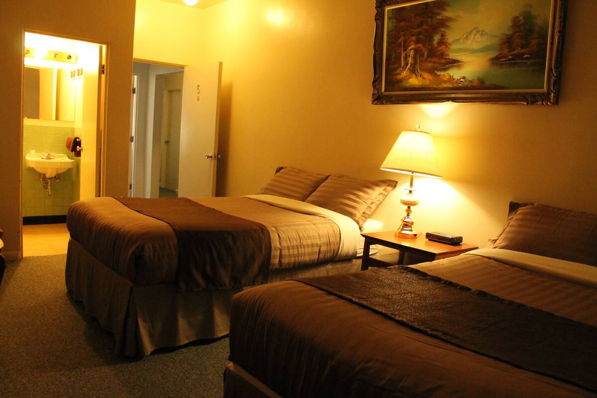 Habitación de hotel con dos camas, una mesita de noche con lámpara, un cuadro en la pared y un baño visible a través de una puerta abierta.