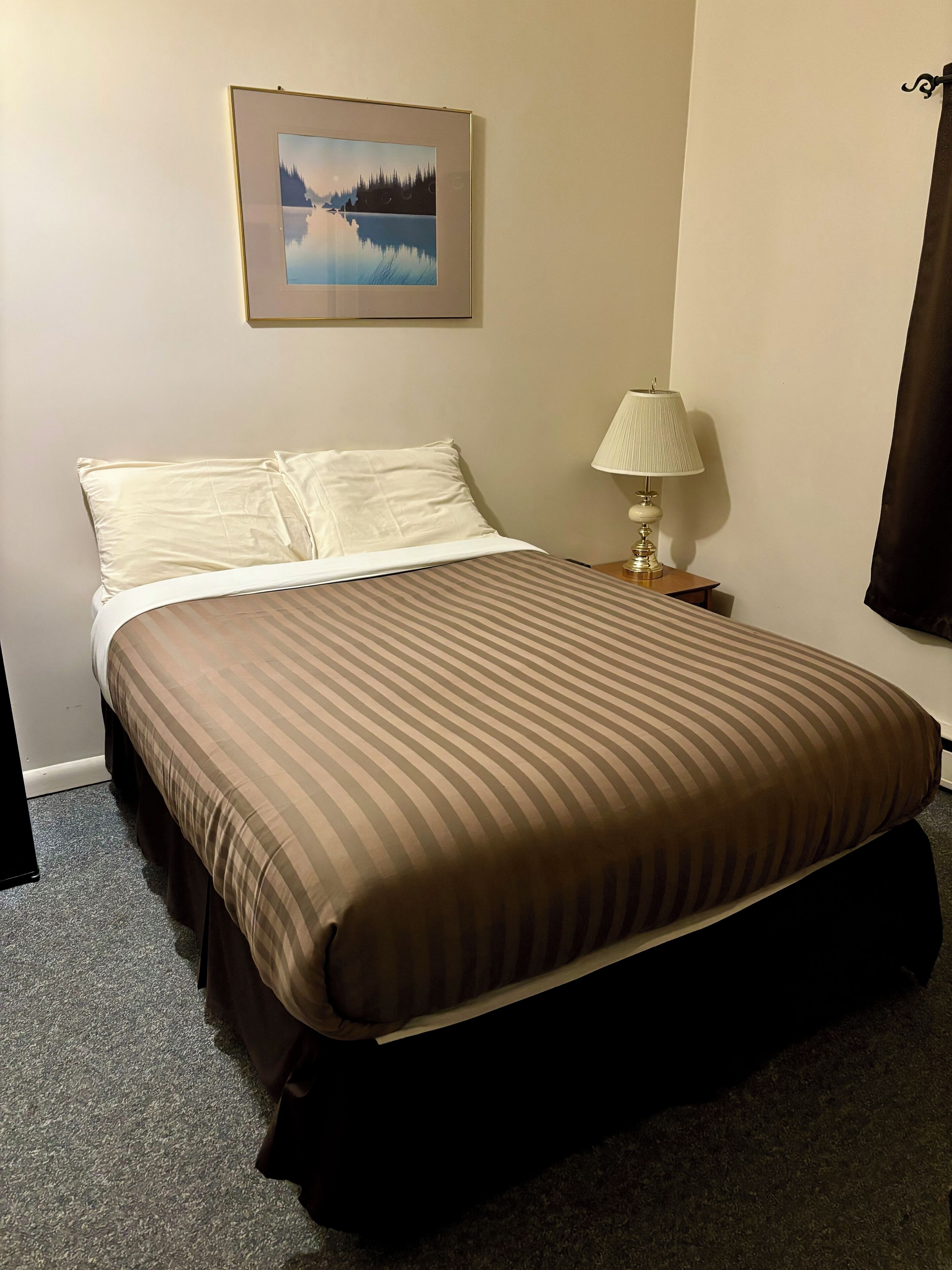 A neatly made bed with a striped bedspread and white pillows, a bedside lamp, and a framed picture of a lake on the wall.
