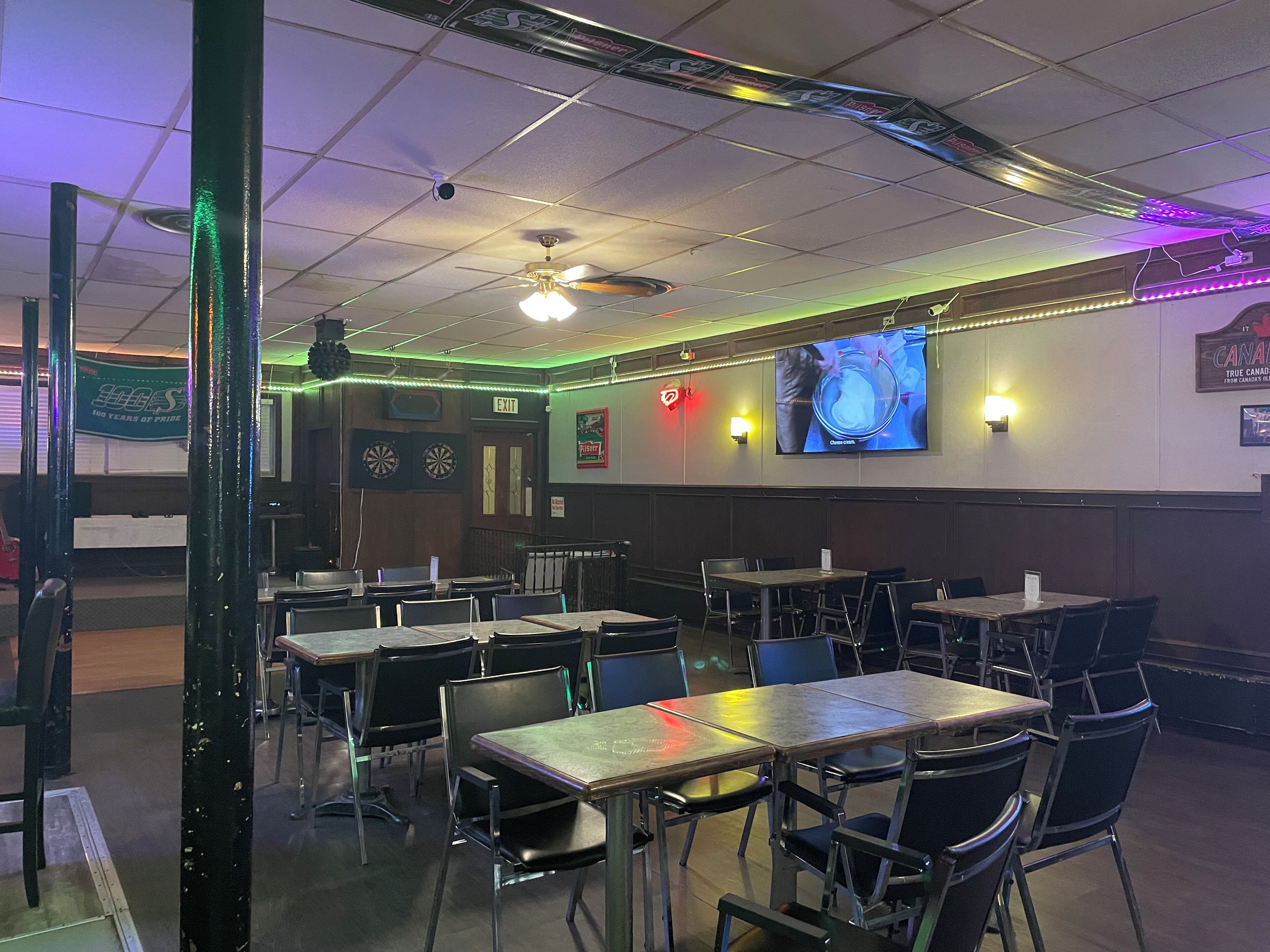 A restaurant interior with tables and chairs, a ceiling fan, dartboards, and a TV screen.
