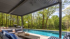 Ein überdachter Außenpoolbereich mit Blick auf einen Wald, ausgestattet mit Sofas und einem Tisch.