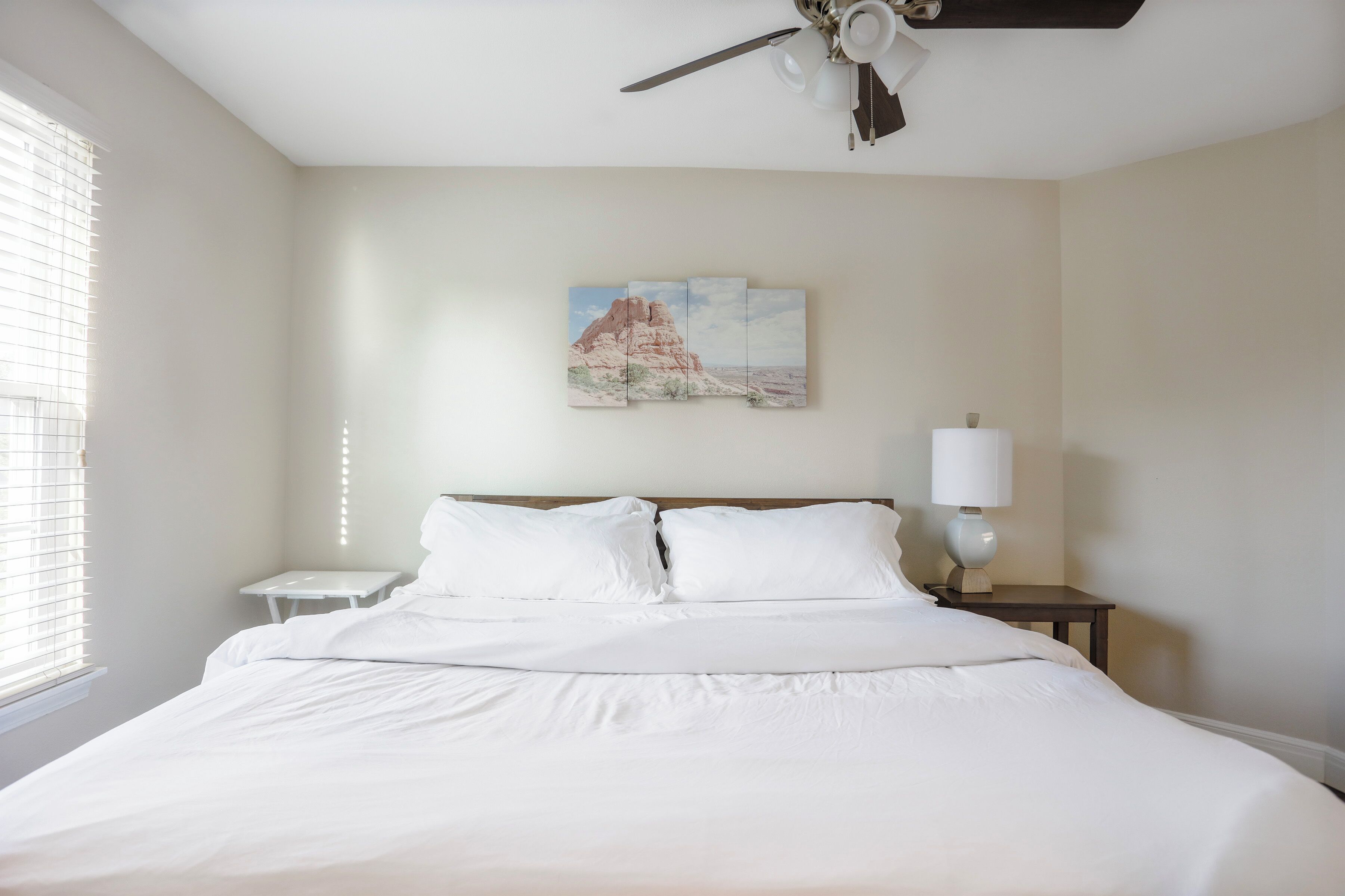 Una cama bien hecha con sábanas blancas, un ventilador de techo, una mesita de noche con una lámpara y un cuadro de un paisaje desértico en la pared.
