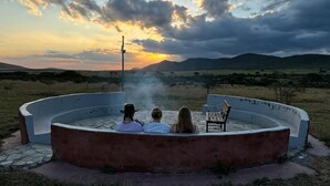A circular outdoor hot tub with three people sitting around it, surrounded by a grassy landscape and hills in the background. - Endeem Mara Resort (Maasai Mara)