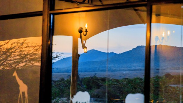A room with a large window offering a view of a savannah landscape with silhouettes of giraffes and elephants. - Endeem Mara Resort (Maasai Mara)