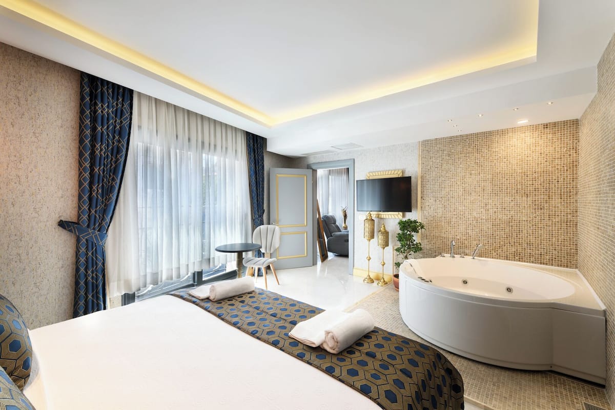 A modern hotel room with a large bed, a separate seating area, a large bathtub, and a flat-screen TV.