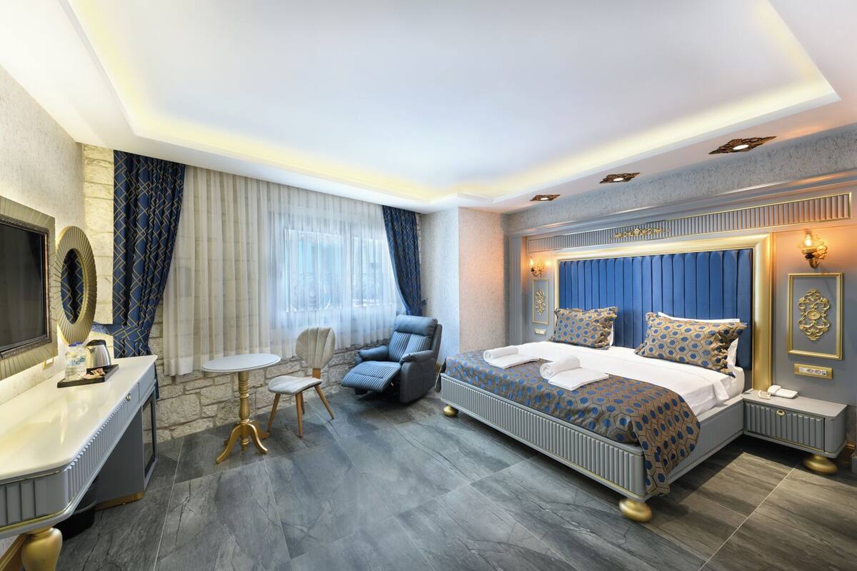 A modern hotel room with a large bed, a seating area with a chair and table, a vanity with a mirror, and a television.