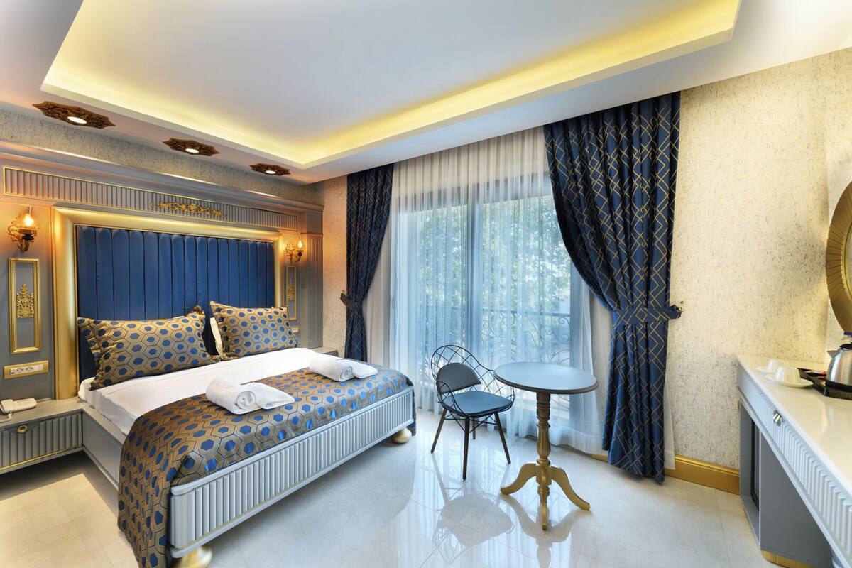 A modern hotel bedroom with a large bed, a bedside table, a chair, and a view of the outdoors.