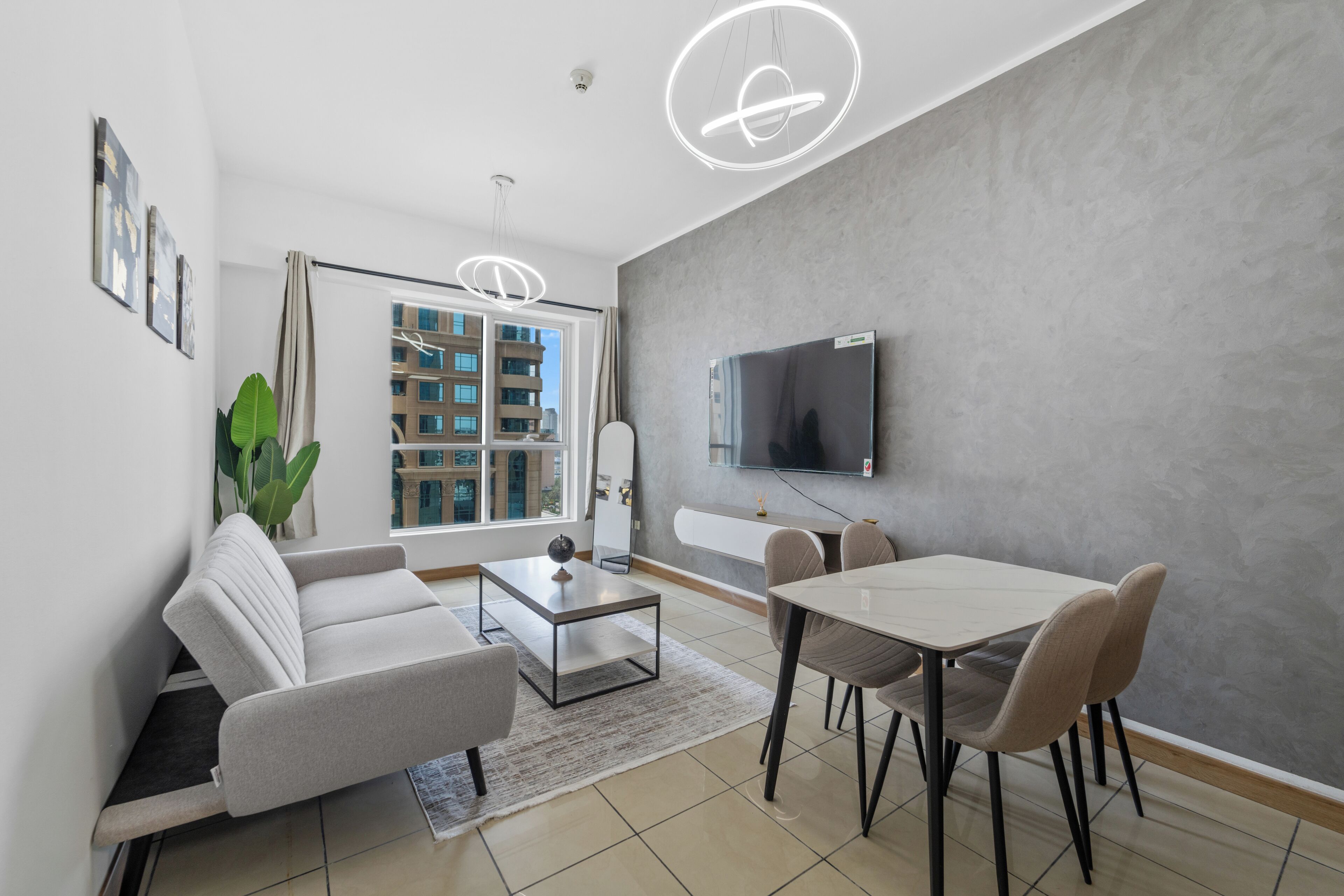 Een moderne woonkamer met een bank, salontafel, eettafel en stoelen. Er is een televisie aan de muur bevestigd en een groot raam met gordijnen.