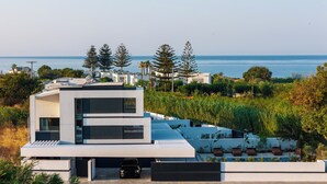 A modern building with a pool and outdoor seating, overlooking a coastal area with trees and a clear sky. - Nuera II: Modern 4BR Villa, 300m from the Beach (Chania)