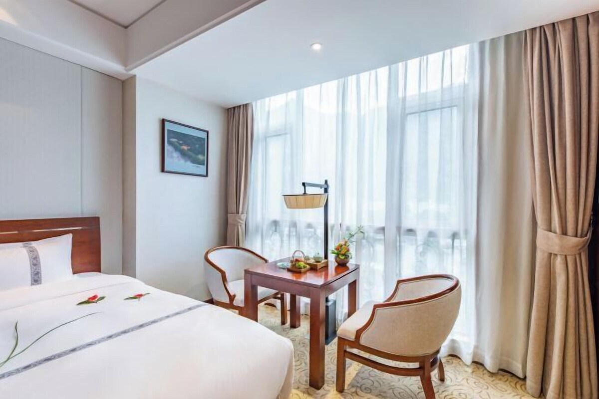 A hotel room with a large bed, a desk with a lamp, two chairs, and a view of the outdoors through sheer curtains.
