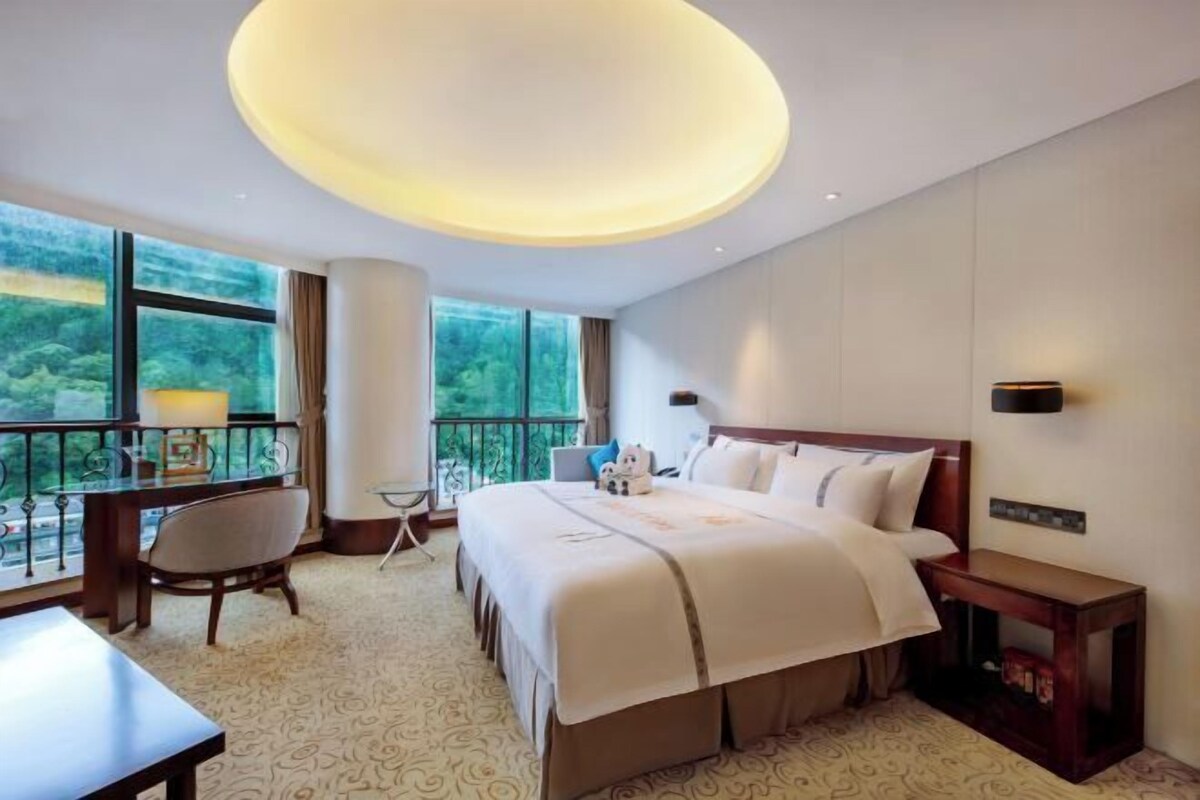 A hotel room with a large bed, a desk, a chair, and a view of greenery through the windows.