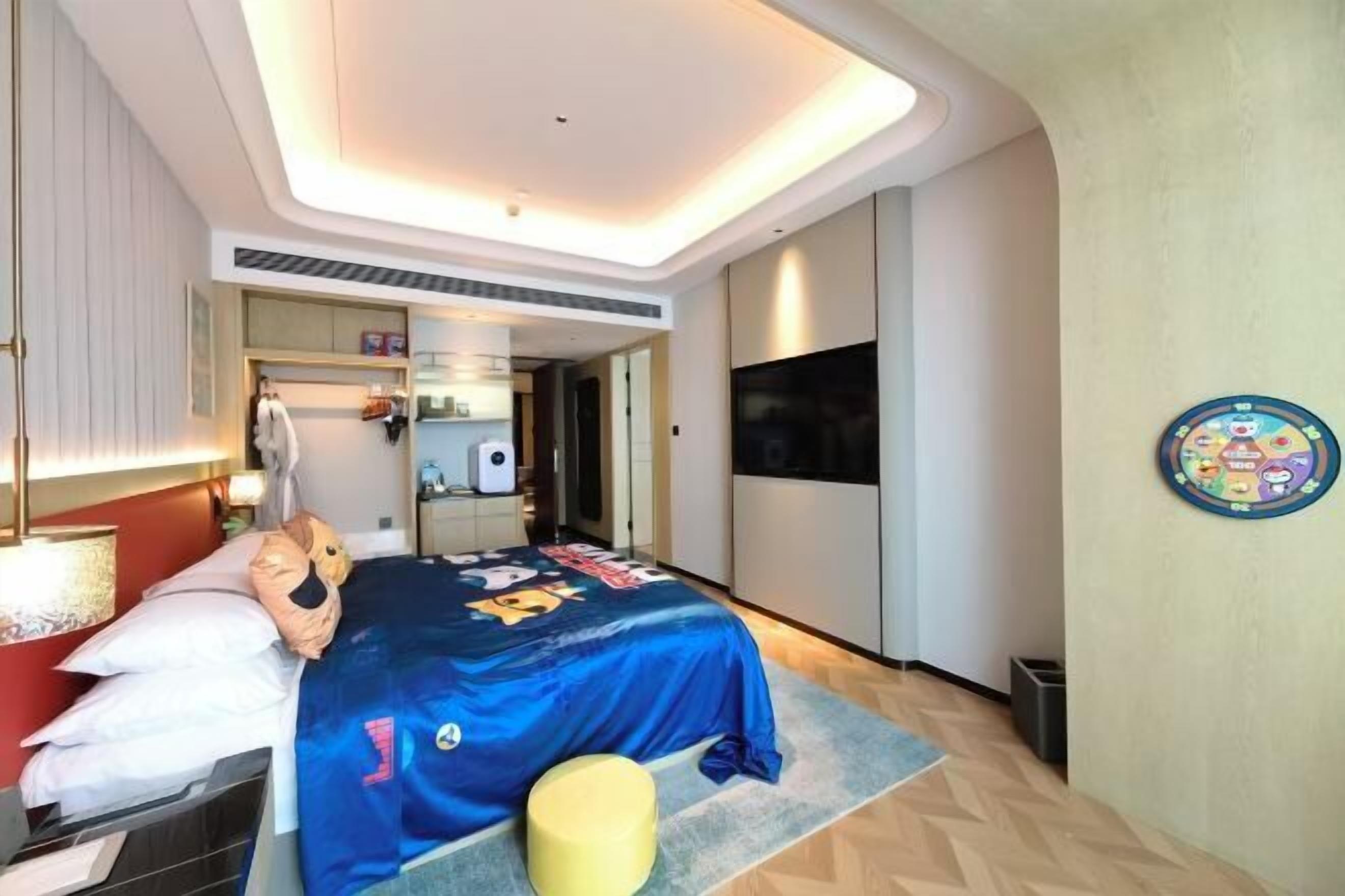 A modern hotel room with a bed, a TV, a circular wall decoration, and a small stool.