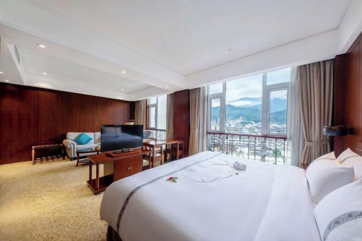 A hotel room with a large bed, a TV, a desk, and a balcony with a city view.