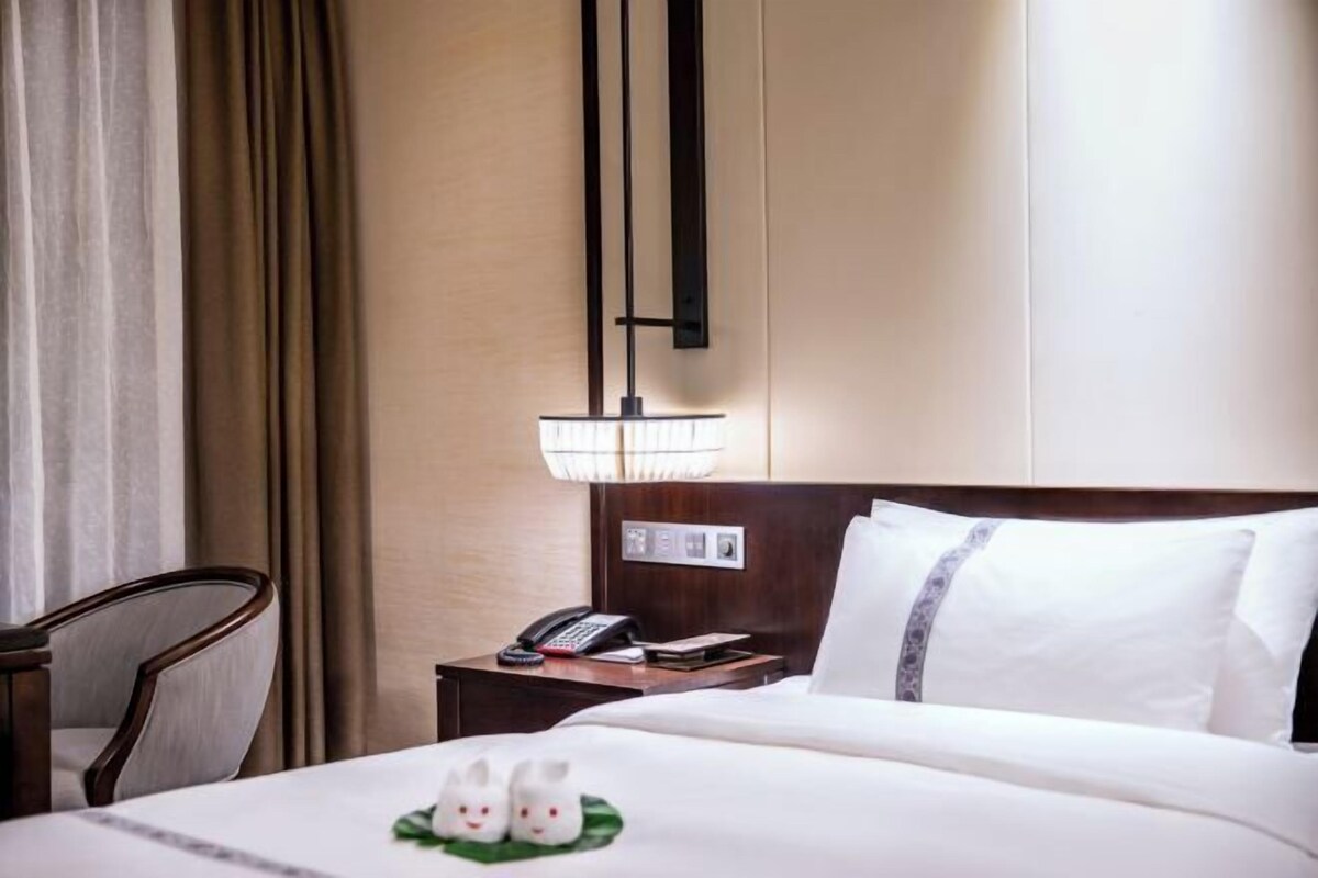 A hotel room with a bed, a nightstand with a lamp, a chair, and a window with curtains.