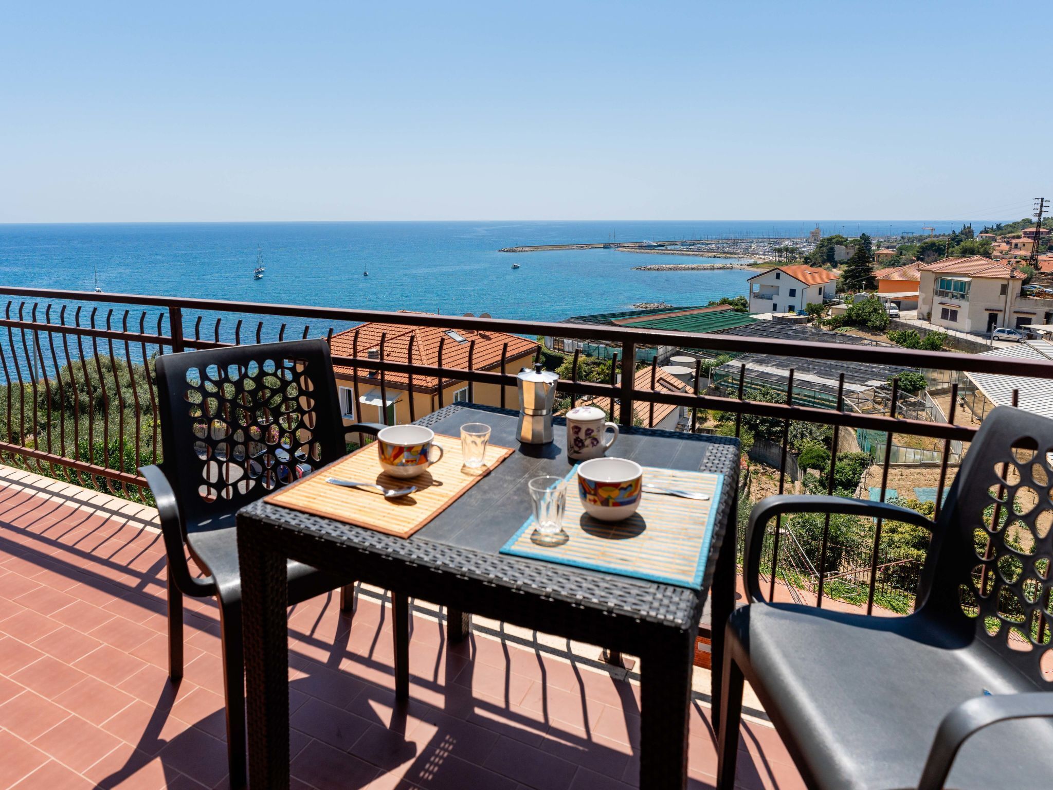 A balcony with a table set for two, overlooking a coastal view with buildings and the sea.