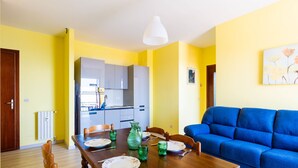 A dining area with a wooden table set for four, a blue sofa, and a kitchen in the background. - Sunny Home by Interhome (Santo Stefano al Mare)