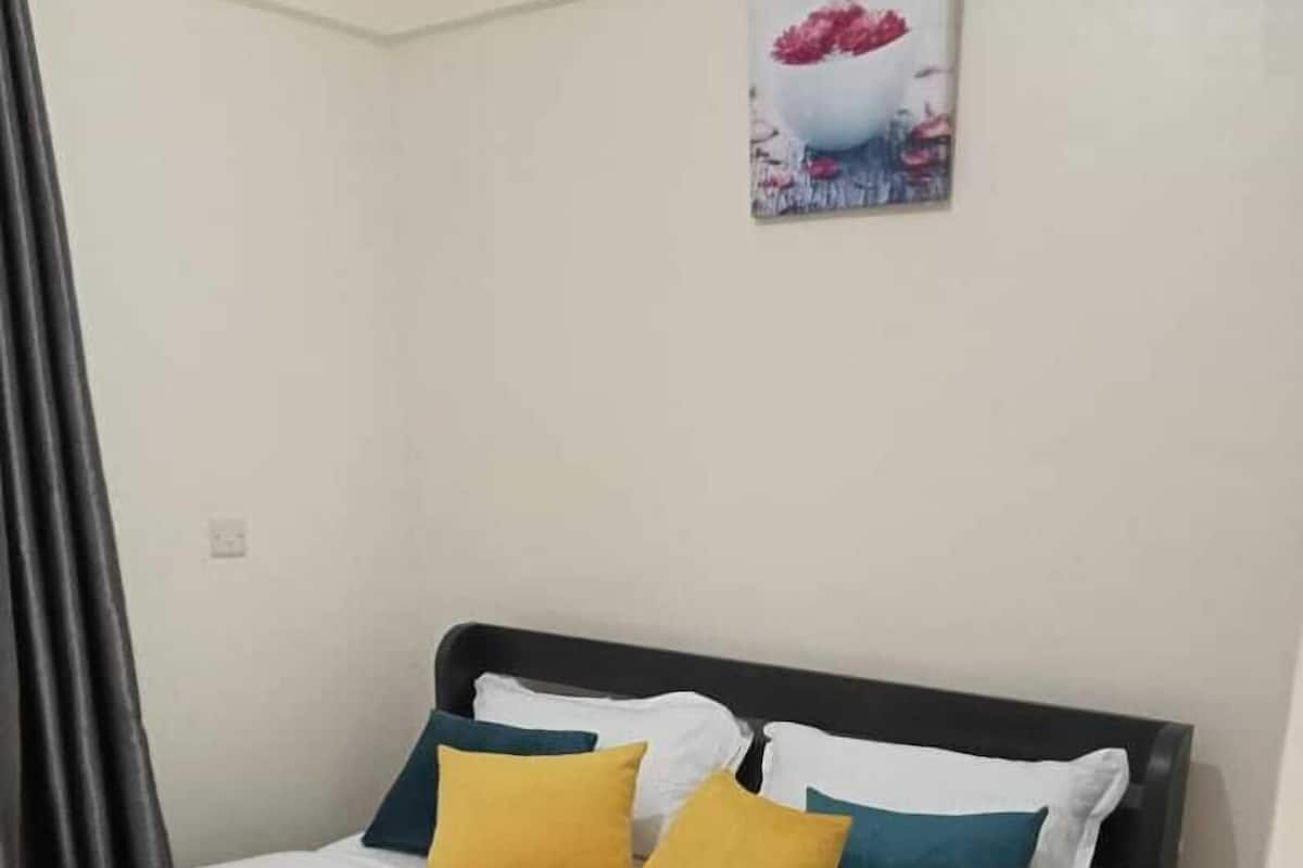 A neatly made bed with white bedding and two pillows, one yellow and one blue, in a room with a plain white wall and a painting above the bed.