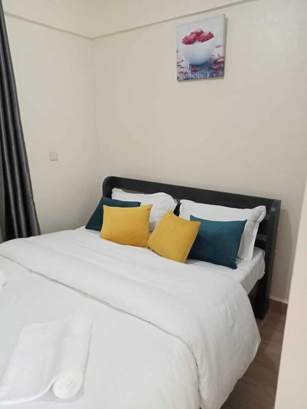 Una cama bien hecha con ropa de cama blanca y dos almohadas, una amarilla y otra azul, en una habitación con una pared blanca lisa y un cuadro sobre la cama.