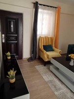 A living room with a dark wooden door, a yellow armchair with a blue cushion, a black coffee table, and a small plant on the side table.