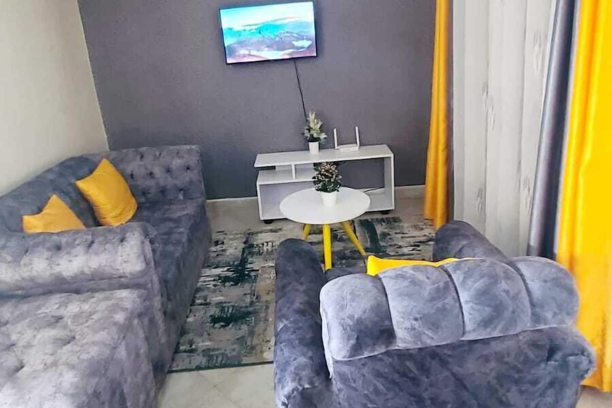 A modern living room with a grey sofa, a smaller grey armchair, a white coffee table with yellow legs, and a flat-screen TV mounted on the wall.